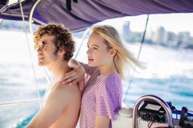 Kız arkadaşıyla bir yatın dümenini tutan ve uzaklara bakan bir adam.