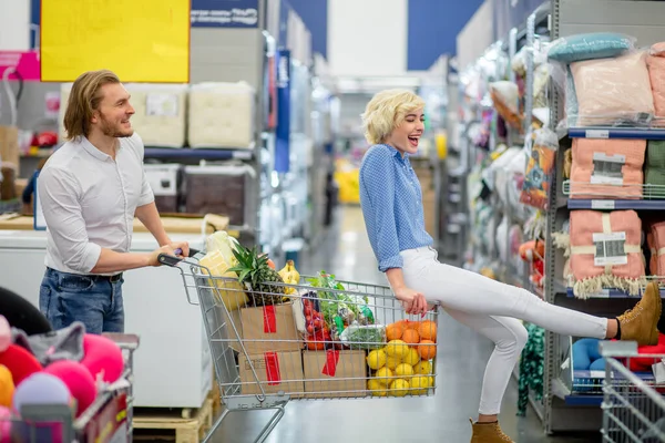 Young happy man pushing shopping trolley with his girfriend in mall — Stock Photo, Image