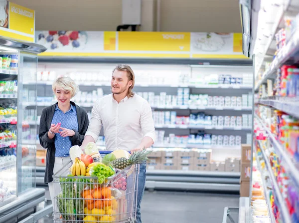 Compradores masculinos e femininos empurram carrinho com produtos de supermercado no corredor do supermercado — Fotografia de Stock
