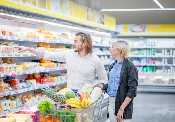 Compradores masculinos e femininos empurram carrinho com produtos de supermercado no corredor do supermercado — Fotografia de Stock