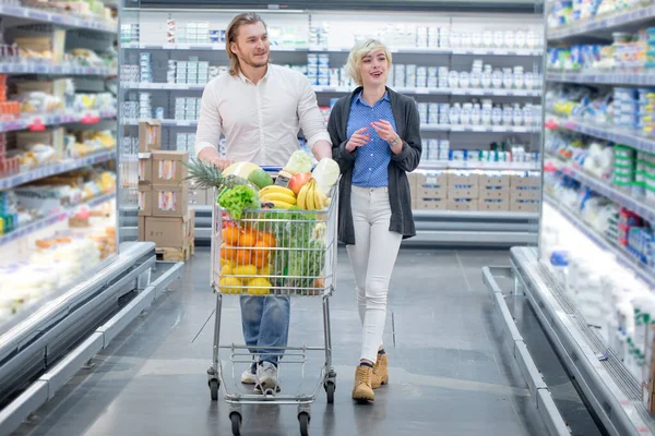 male and female shoppers push cart with grocery products in supermarket aisle