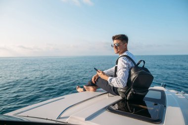 Güneş gözlüklü bir adam yat kullanıyor ve telefonda konuşuyor.