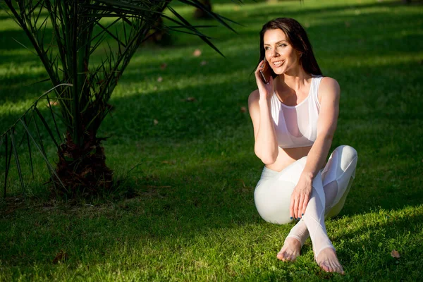 Chica de yoga sentado en la hierba, hablando en el teléfono inteligente en el parque —  Fotos de Stock
