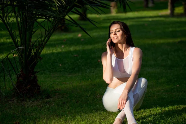 Jóga dívka sedí na trávě, mluví na smartphone v parku — Stock fotografie