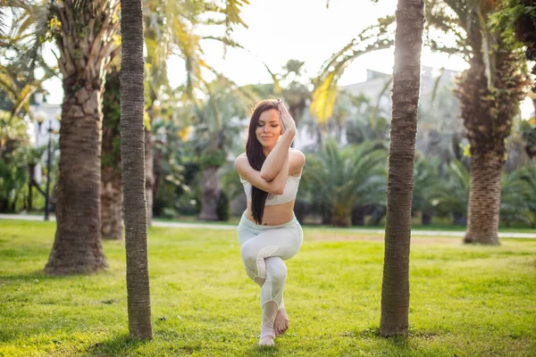 Kvinna i vit sport outfit utför Yoga Eagle pose utomhus, i parken äng. — Stockfoto