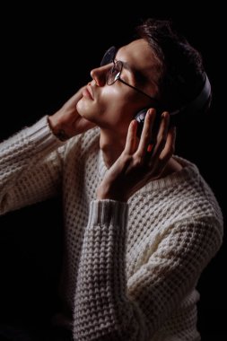 Gözlüklü ve kulaklıklı genç adam siyah arka planda müzik dinliyor.