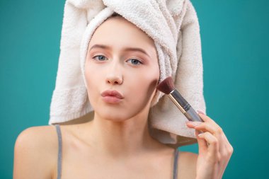 Duştan sonra kafasına havlu geçirmiş bir kadın. Elinde kilden maske takmak için fırça tutuyor.