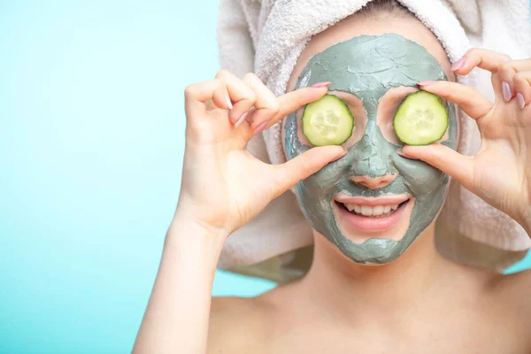 Wanita muda dengan masker wajah dan irisan mentimun terisolasi di atas dinding biru studio — Stok Foto