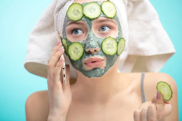 Woman with towel on head and face mask enriching with cucumbers via smartphone — Stock Photo, Image