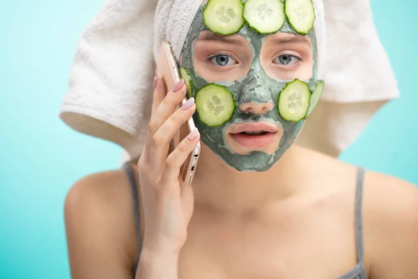 Woman with towel on head and face mask enriching with cucumbers via smartphone — Stock Photo, Image