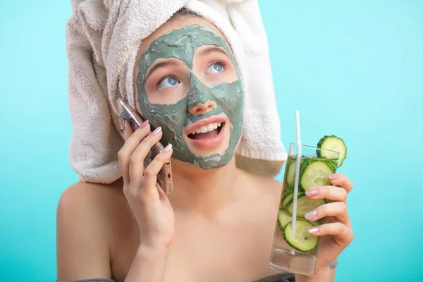 Vrouw met handdoek op hoofd en gezichtsmasker verrijkt met komkommers via smartphone — Stockfoto