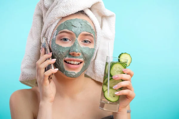 Žena s ručníkem na hlavě a obličejovou maskou obohacující okurky přes smartphone — Stock fotografie