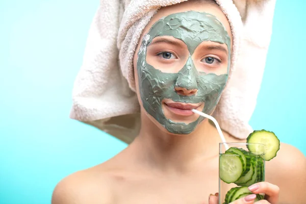 Mujer con máscara de arcilla cosmética beber agua cucucmber aislado en el estudio azul — Foto de Stock