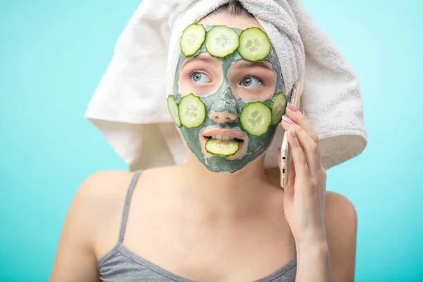 Mujer con toalla en la cabeza y mascarilla enriquecedora con pepinos a través de smartphone — Foto de Stock