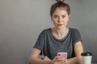 Saçları topuzlu sarışın bir kadın gri üzerinde akıllı bir telefon tutuyor.