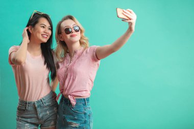 Mutlu genç kızlar mavi duvara karşı akıllı telefonlarıyla selfie çekiyorlar.