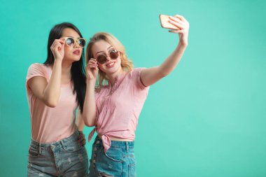 Mutlu genç kızlar mavi duvara karşı akıllı telefonlarıyla selfie çekiyorlar.
