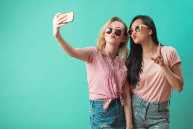 Mutlu genç kızlar mavi duvara karşı akıllı telefonlarıyla selfie çekiyorlar.