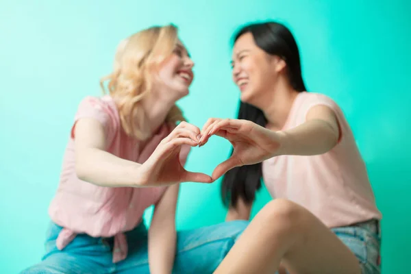 Chicas jóvenes mostrando el signo del corazón con las manos sentadas sobre fondo azul — Foto de Stock