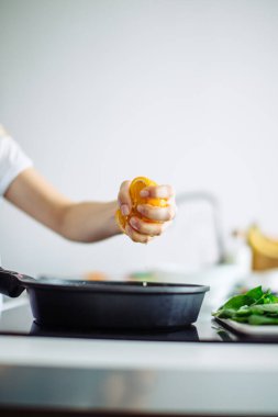 Genç ev hanımı mutfakta yemek pişiriyor, tavaya portakal sıkıyor.