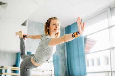 Genç, formda bir kadın yoga pozu veriyor. Bir bacağı havada duruyor..