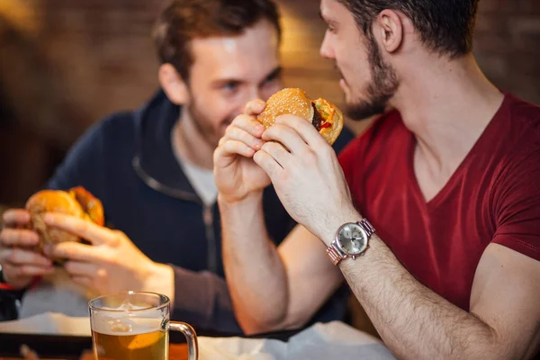 Two happy male friends eating tasty burgers in bar.