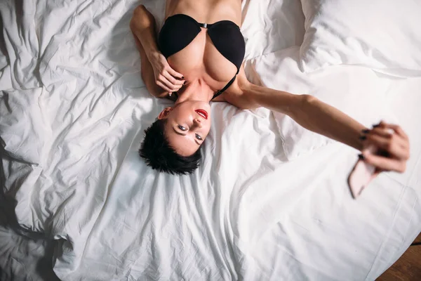 Aantrekkelijke sexy brunette vrouw ligt op bed in lingerie en het nemen van selfie op telefoon. — Stockfoto