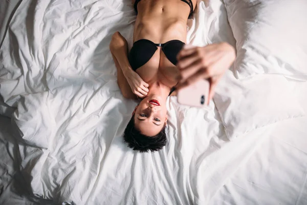 Attraente sexy bruna donna si trova su letto in lingerie e prendendo selfie su telefono. — Foto Stock