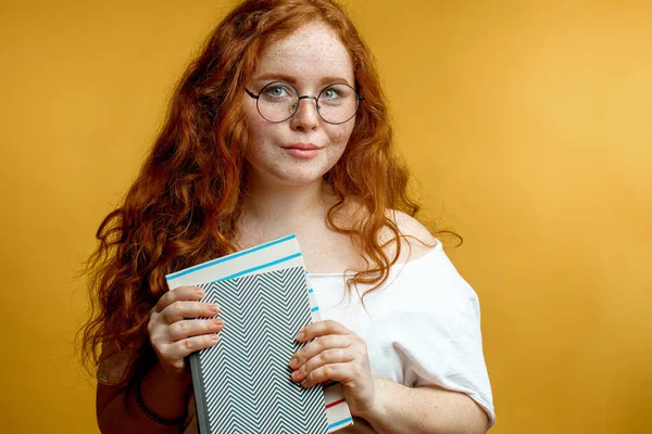 Ruiva jovem segurando livros em branco olhando para a câmera isolada amarelo — Fotografia de Stock