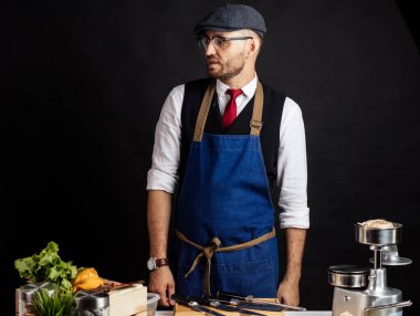 Aşçı stüdyoda, siyah duvara karşı tahta bir masanın yanında duruyor..