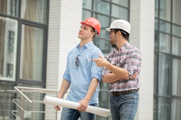 Beau constructeur tient le projet tandis que son collègue pointant vers le bâtiment — Photo