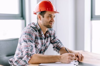 Neşeli inşaat işçisi elinde kalem ve kağıtla oturuyor.