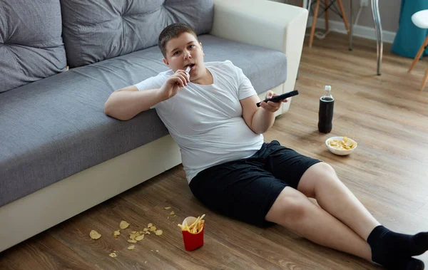 Sobrepeso chico gordo comer comida chatarra mientras viendo tv solo en casa —  Fotos de Stock