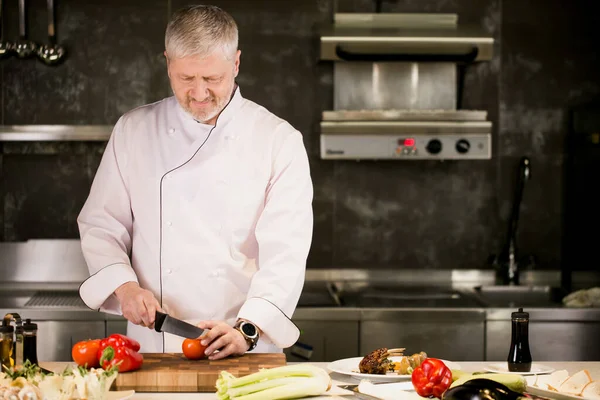 Hombre mayor con barba en uniforme blanco está cortando verduras en la cocina — Foto de Stock