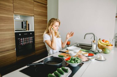 Genç bir kadın mutfakta yemek pişirirken cep telefonu kullanıyor.