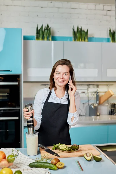 Chica positiva preparando el desayuno y tener una conversación por teléfono en la cocina — Foto de Stock