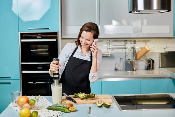 Sorrindo menina bonita está misturando milkshake ao falar no telefone inteligente — Fotografia de Stock