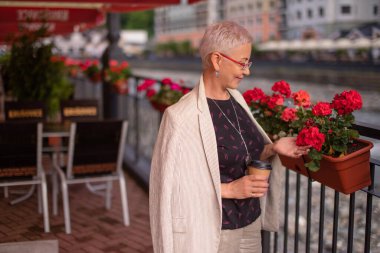 Olgun, gülümseyen bir kadın saksıdaki kırmızı çiçeklerin tadını çıkarıyor.