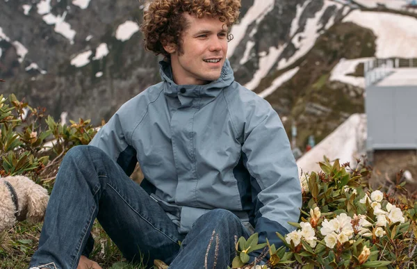 Touristen-Hipster-Mann sitzt auf dem Gras mit Blumen. ein schneebedeckter Berg — Stockfoto