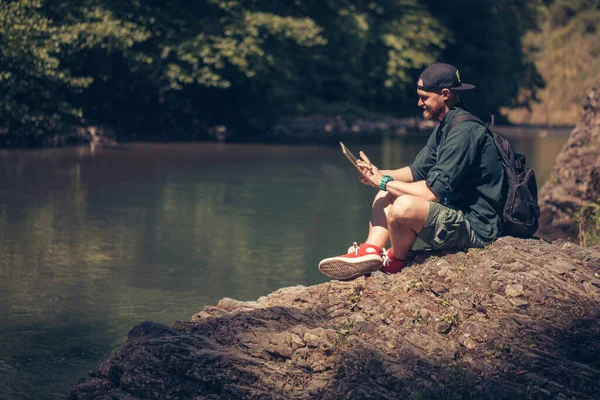 Одинокий чоловічий пішохід, що використовує планшет на природі, сидячи на скелястому березі річки — стокове фото