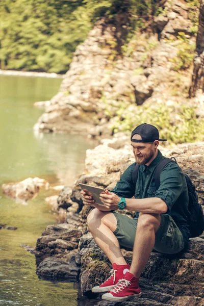 Jeden muž turista pomocí tabletu na přírodě, zatímco sedí na skalnatém břehu řeky — Stock fotografie