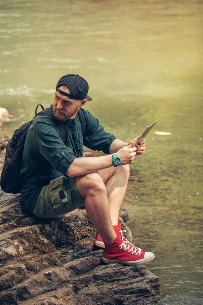 Ενιαία αρσενικό πεζοπόρος χρησιμοποιώντας tablet στη φύση, ενώ κάθεται σε βραχώδη όχθη του ποταμού — Φωτογραφία Αρχείου
