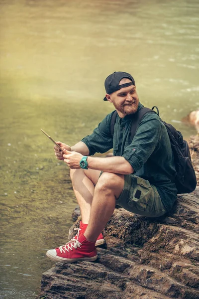 Jeden muž turista pomocí tabletu na přírodě, zatímco sedí na skalnatém břehu řeky — Stock fotografie