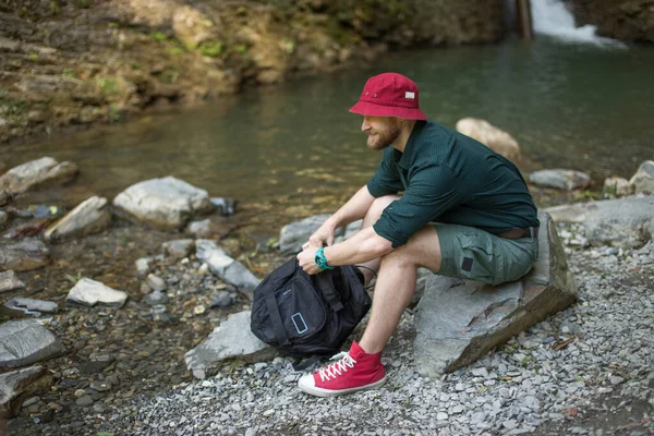 Ein Tourist mit Rucksack sitzt in der Nähe eines Sees in einem wilden Sommerwald. Expedition — Stockfoto