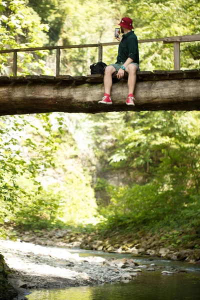 O viajante senta-se na ponte de madeira e usa o smartphone para ver o mapa para navegar. — Fotografia de Stock