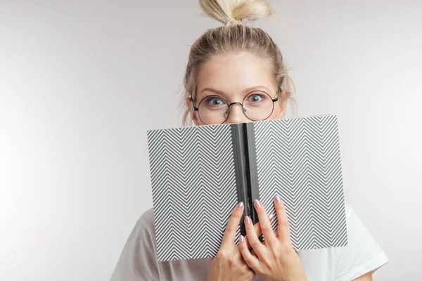 Şok olmuş gözlü sarışın bir kadın açık bir kitabın arkasına saklanıp kameraya bakıyor.. — Stok fotoğraf