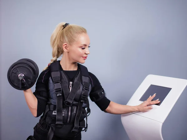 Femme blonde en costume de stimulation musculaire électrique debout avec haltère — Photo