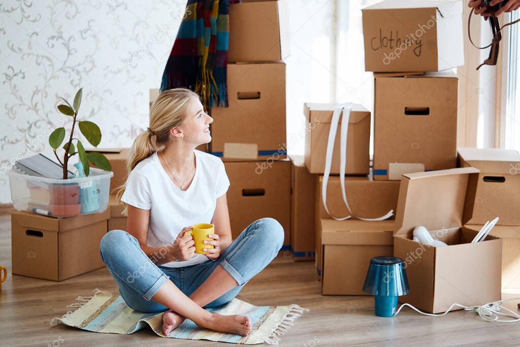 woman with tea in hand sitting on floor of new apartment, pile of moving boxes on background