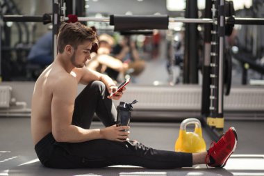 Sportif erkek sporcu spor salonunda elinde su şişesiyle akıllı telefonuna bakıyor..