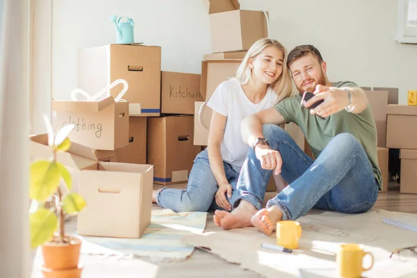 Pasangan pindah di rumah baru. Duduk di lantai dan bersantai setelah unpacking.Doing selfie dengan smartphone — Stok Foto